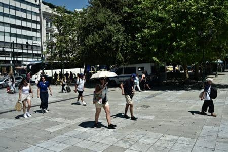 Άνθρωποι περπατούν σε αστικό περιβάλλον στην Αθήνα, με έναν να χρησιμοποιεί ομπρέλα για προστασία από τον ήλιο κατά τη διάρκεια καύσωνα.