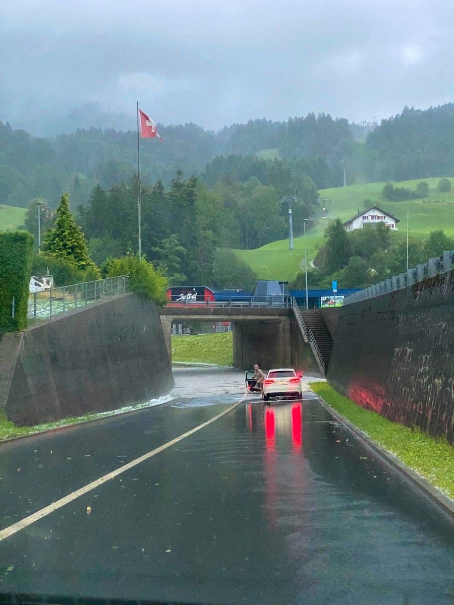 A car partially submerged in a heavily flooded underpass in a Swiss town, surrounded by green hills and a Swiss flag.