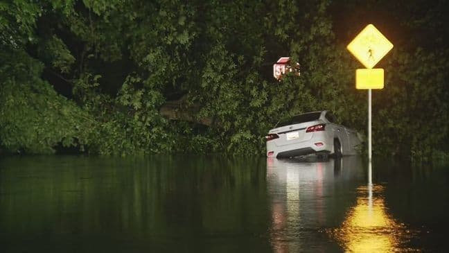 A car partially submerged in dark floodwaters on a road at night, with a pedestrian crossing sign nearby, depicting a flash flood impacting commuters.