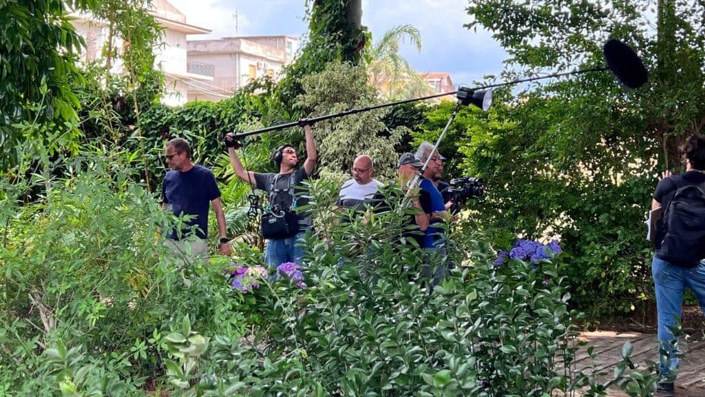 A film crew works amidst lush greenery, symbolizing Milazzo's fertile hinterlands and efforts to rediscover forgotten agricultural treasures.
