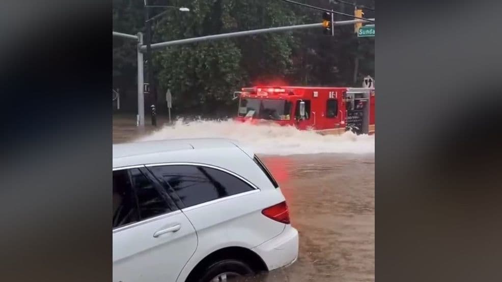 A fire truck drives through a deeply flooded urban street, splashing water, with cars partially submerged, illustrating severe urban flash flooding.