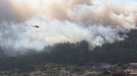 A firefighting helicopter battling a large wildfire engulfing forested hills and threatening homes in Ponteceso, illustrating the widespread devastation.