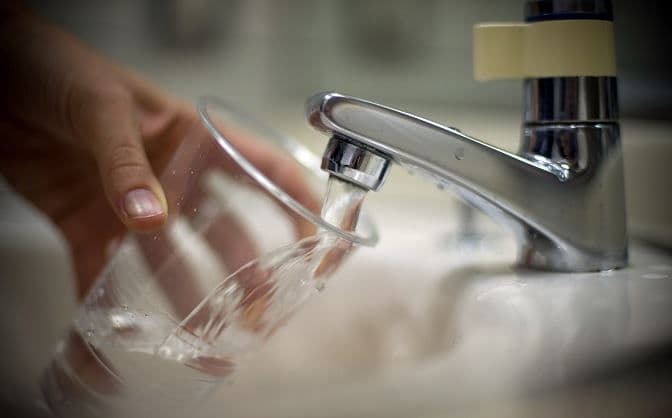 A hand holding a glass under a running water tap, illustrating daily potable water supply.