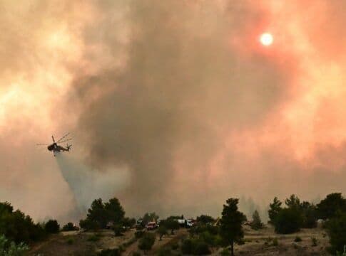 A helicopter drops water on a large wildfire with thick smoke and a hazy red sun, highlighting extreme weather impacts in Greece.