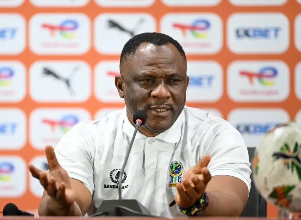 A male football coach speaking at a press conference, with a microphone and football visible, representing insights from CHAN 2025 technical benches.