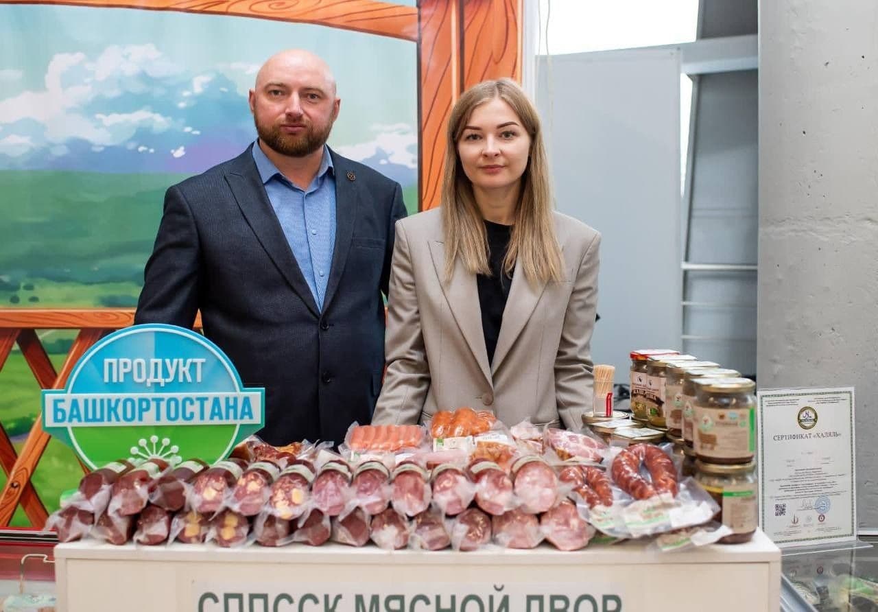 A man and a woman proudly standing behind a display of packaged meat products, featuring the 'Product of Bashkortostan' label.