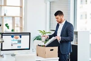 A man in a suit packing personal items into a box at his office desk, symbolizing job loss and the vulnerability of workers under closed permits.