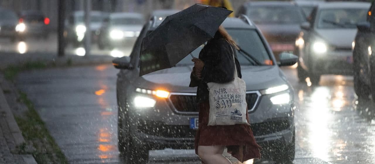 A pedestrian crosses a street in Bautzen, Saxony, during heavy rainfall, holding an umbrella, with cars on the wet road. This image illustrates the severe storm alert and the 'coming deluge' in Germany.