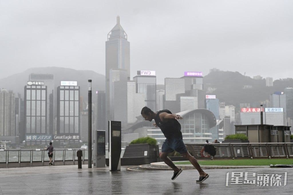 A person leans against strong winds on a waterfront promenade with the Hong Kong skyline in the background during a storm, illustrating urban resilience.