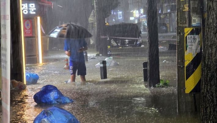 A person with an umbrella wading through a heavily flooded street in a South Korean city during extreme rainfall.