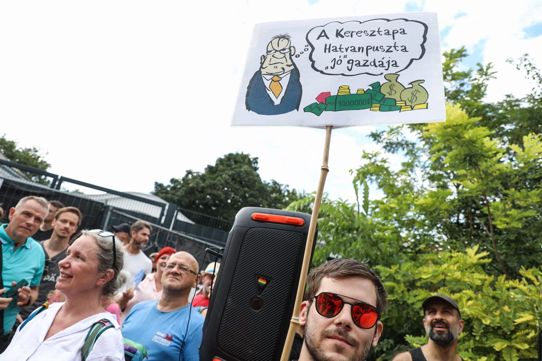 A protester holding a sign depicting a cartoon figure with money bags, questioning wealth at the Hatvanpuszta transparency demonstration.