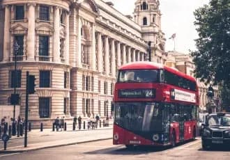 A red double-decker bus driving past historic buildings in a European city, representing urban life and landscapes shaped by climate.
