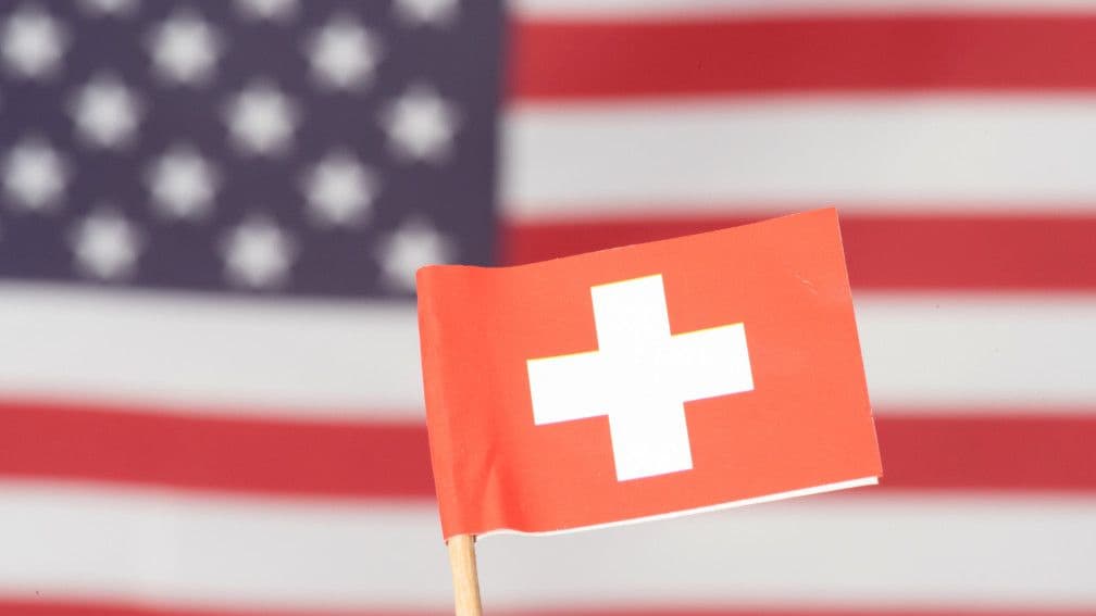 A small Swiss flag in front of a blurred US flag, symbolizing the strained diplomatic relationship between the two nations.