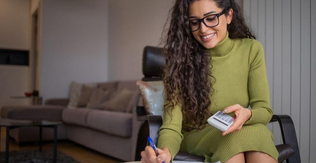 A smiling woman using a calculator and writing, representing a taxpayer navigating their income declaration.