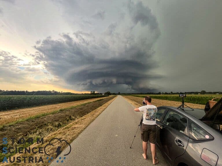 A storm chaser capturing a massive, rotating supercell thunderstorm over a rural Austrian landscape.