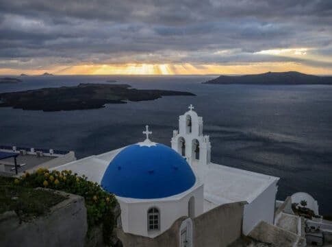 A traditional Greek church overlooking the sea, with sun rays breaking through dramatic clouds, symbolizing Greece's evolving weather.