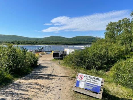 A view of the cross-dam blocking the Tana River at Seida, Norway, surrounded by green banks under a blue sky.