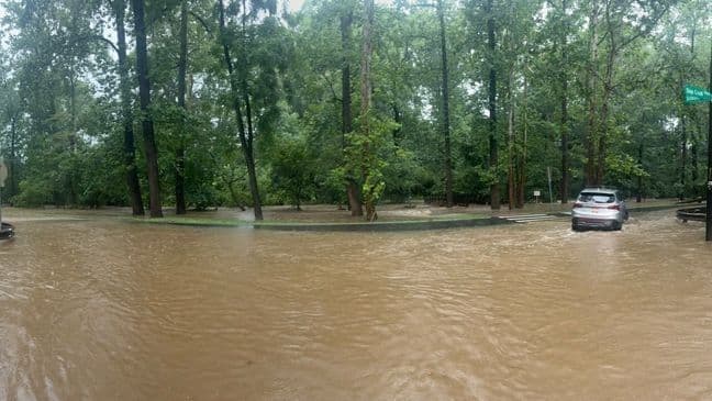 A wide urban road is completely covered in brown floodwater, with cars driving through, illustrating the widespread impact of urban flash flooding on transportation.