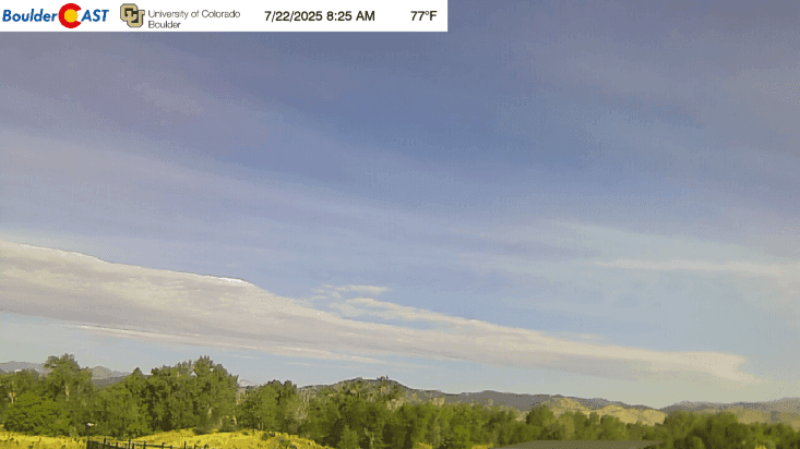 A wide view of a clear sky with wispy clouds over a landscape of green trees and distant mountains under a bright sun.