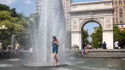A woman stands in a fountain in New York City, seeking relief from extreme heat, symbolizing urban resilience to changing weather.
