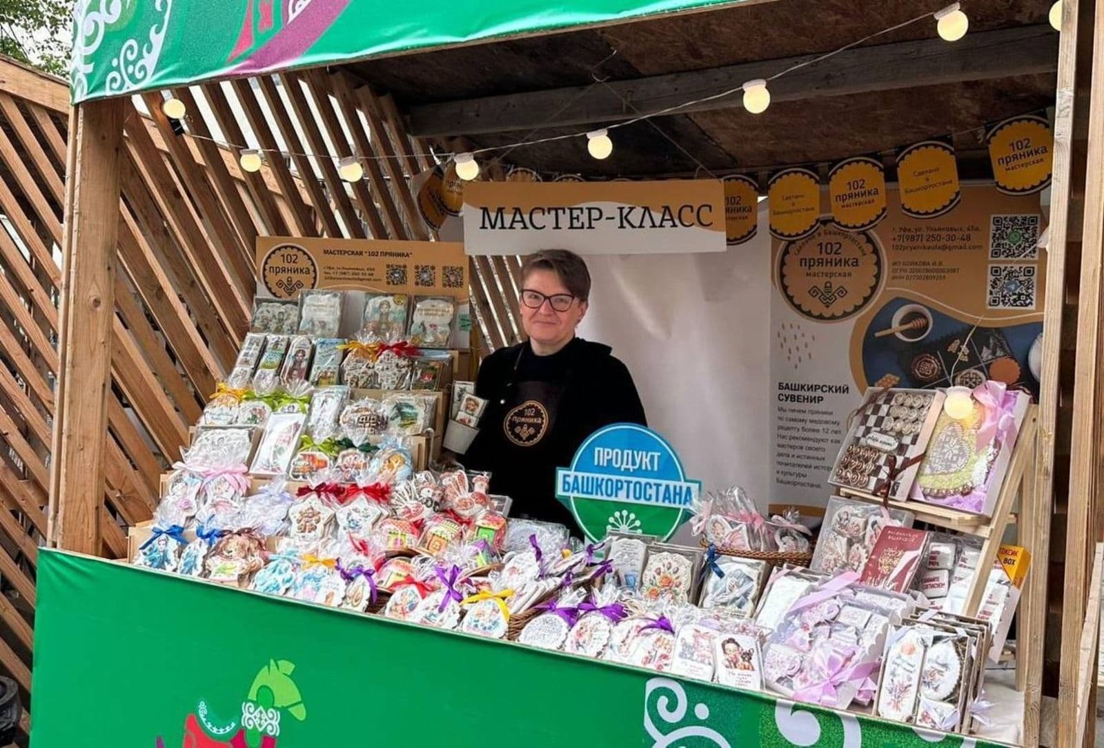 A woman vendor smiling at a vibrant market stall filled with local gingerbread and other products, featuring the 'Product of Bashkortostan' sign.