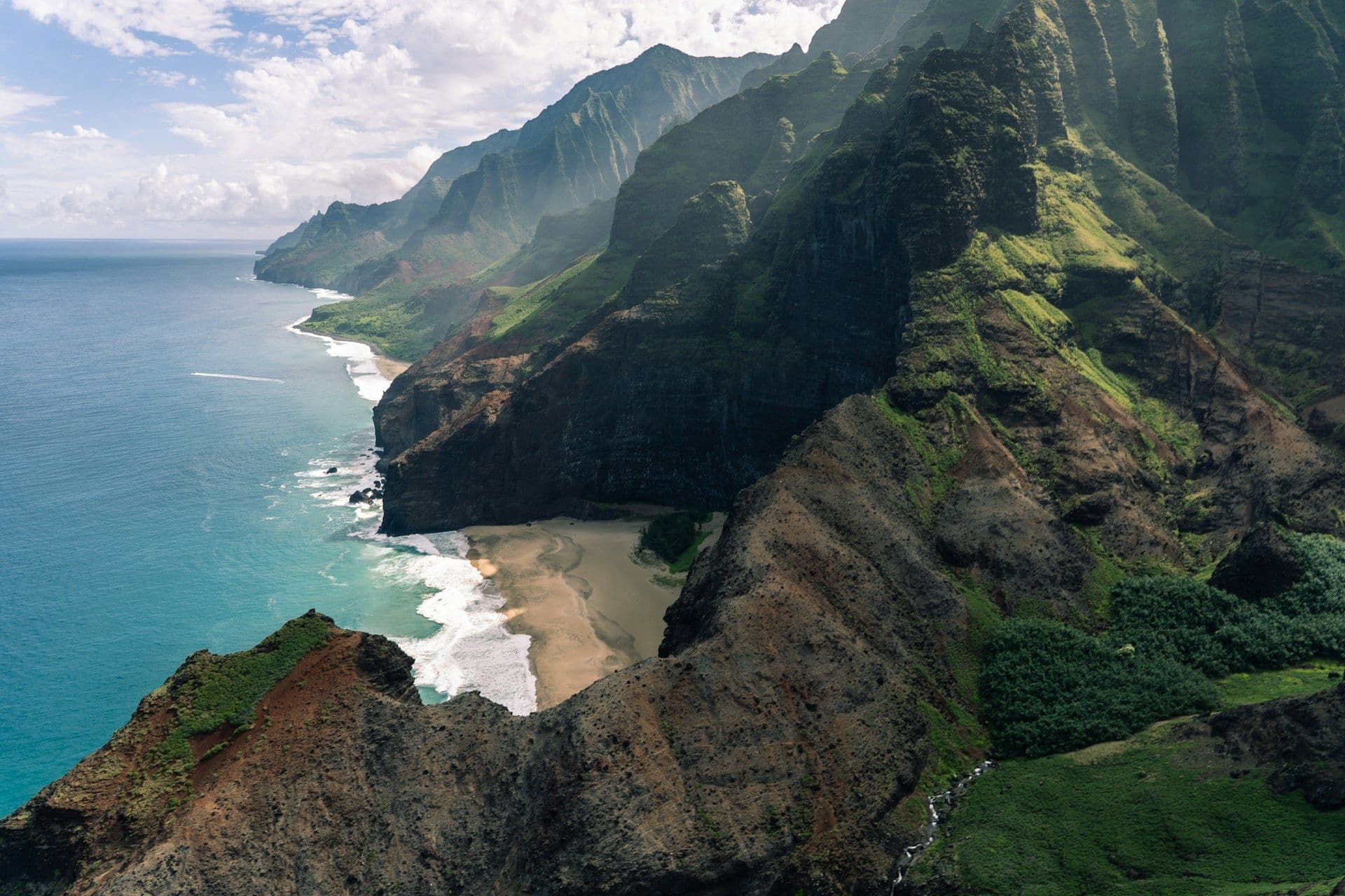 Aerial view of a dramatic green mountain coastline meeting the ocean under a cloudy sky, representing the dynamic forces of weather.