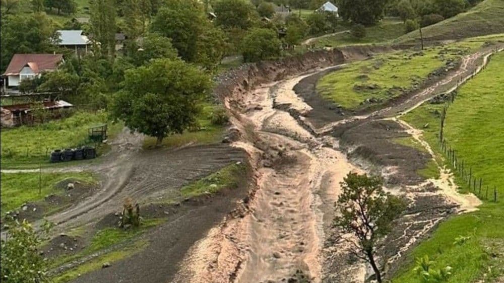 Aerial view of a wide, muddy riverbed or mudslide path winding through green hills with houses, illustrating the impact of heavy mountain rains in Kyrgyzstan.