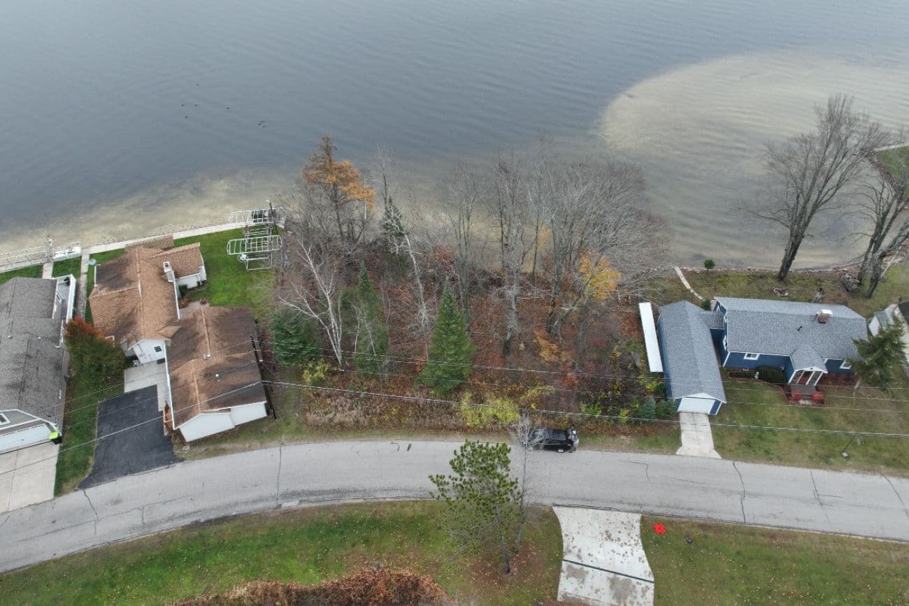 Aerial view of a wooded lakefront parcel of land, bordered by houses and a road, available for auction in Michigan.