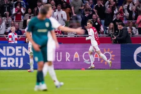 Ajax player in red and white kit celebrating a goal, pointing to the crowd.