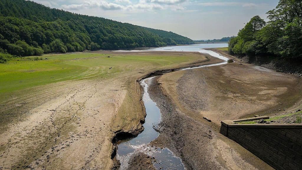 An exposed, cracked reservoir bed with a narrow stream of water, illustrating severe drought conditions and parched earth.