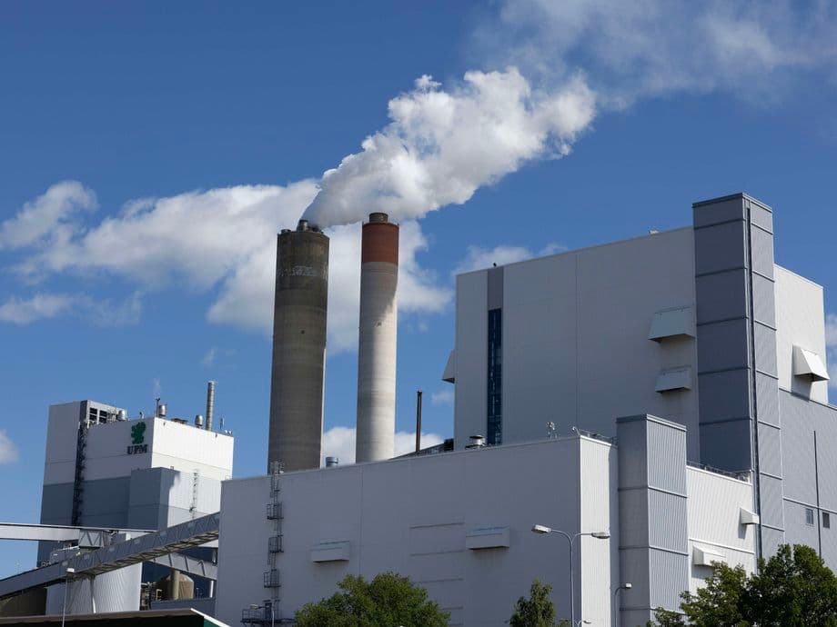 An industrial complex of UPM, featuring large white buildings and two tall smokestacks, one emitting white smoke, under a clear blue sky. The UPM logo is visible on one of the buildings.