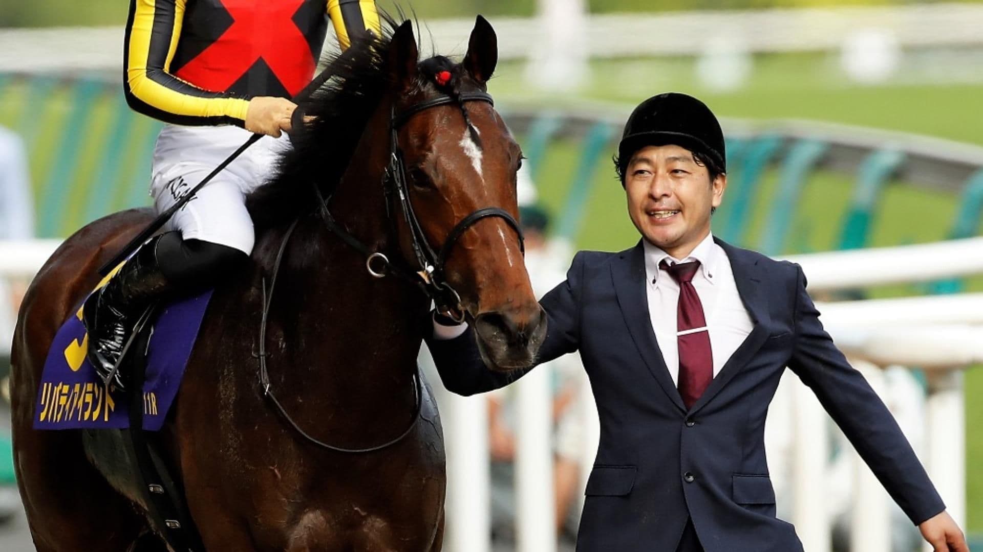 Assistant trainer Keisuke Matsuzaki smiling and walking alongside champion racehorse Liberty Island with a jockey on her back.