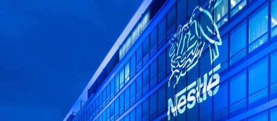 Bâtiment moderne avec le logo Nestlé illuminé en bleu, symbolisant la pression financière sur l'entreprise.