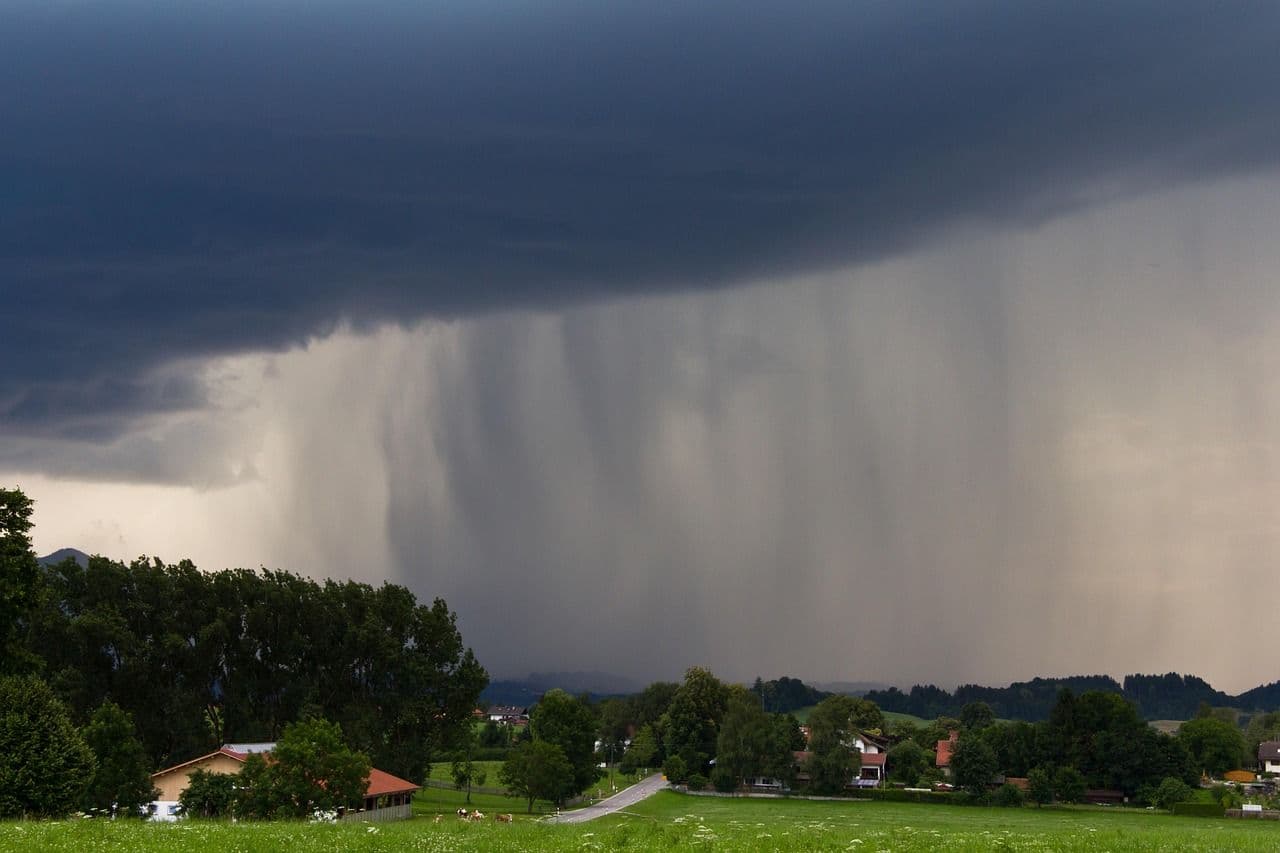 Dark storm clouds and heavy rain falling over a landscape with houses and trees, symbolizing Biysk's rainy August.