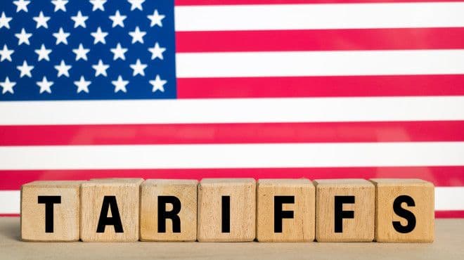 Des blocs de bois épelant 'TARIFFS' devant un drapeau américain, symbolisant les droits de douane imposés par les États-Unis.