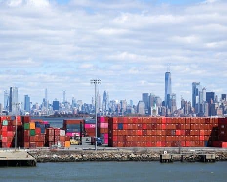 Des conteneurs d'expédition empilés dans un port, avec la skyline de Manhattan en arrière-plan, symbolisant le commerce international et les importations.
