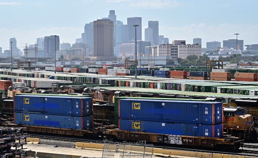 Des conteneurs d'expédition empilés sur des wagons dans un terminal ferroviaire, avec une ville en arrière-plan, illustrant les défis du commerce mondial et des chaînes d'approvisionnement sous l'effet des tarifs.