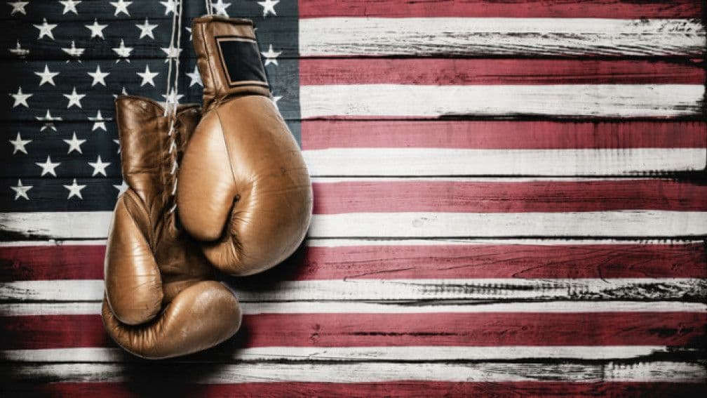 Des gants de boxe suspendus devant un drapeau américain, symbolisant le 'coup de massue' des nouveaux tarifs douaniers.