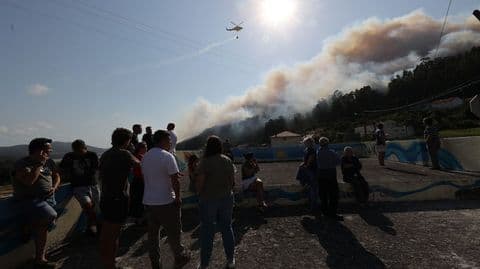 Des habitants de Ponteceso observent un vaste incendie de forêt et un hélicoptère de lutte contre les incendies sous un soleil éclatant.