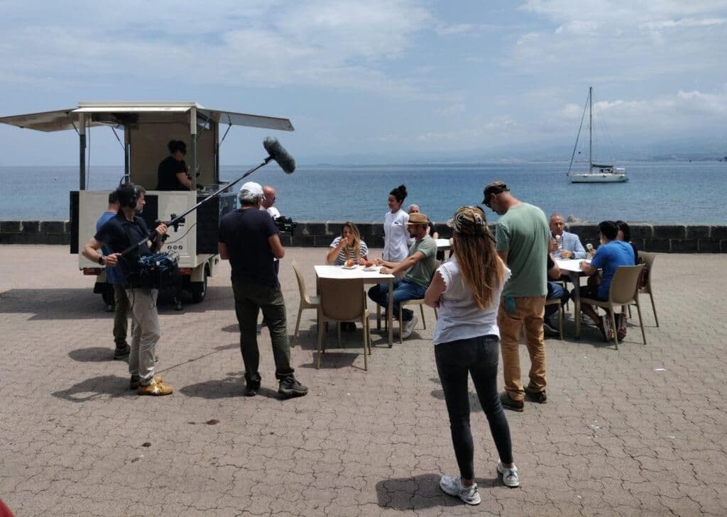 Des personnes dégustant des plats sur une plage de Milazzo, avec un food truck et la mer en arrière-plan, évoquant les saveurs maritimes et l'histoire vivante.