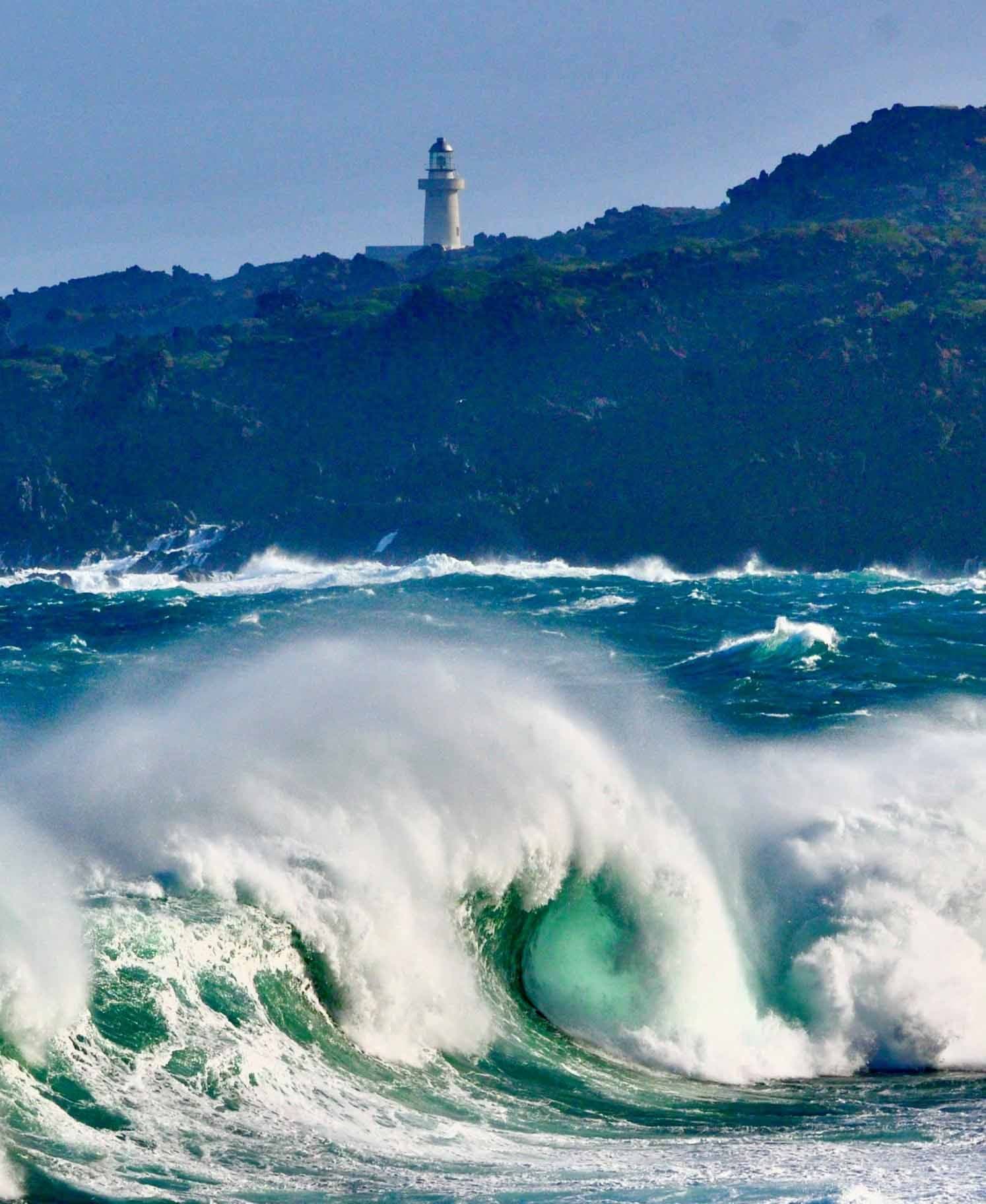 Dramatic large wave crashing near a lighthouse on the rocky coast of Pantelleria, symbolizing the island's dynamic nature and maritime challenges.