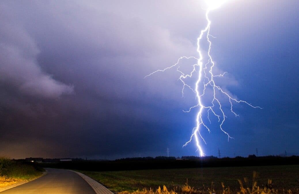 Ein heller Blitz durchschlägt den dunklen Nachthimmel über einer ländlichen Straße in Österreich, symbolisch für die zunehmende Wetterextreme.