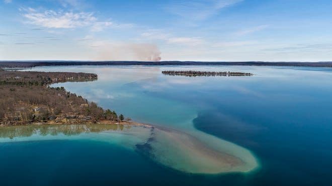 Expansive aerial view of a large Michigan lake with a forested island and distant shoreline, representing untamed natural beauty.