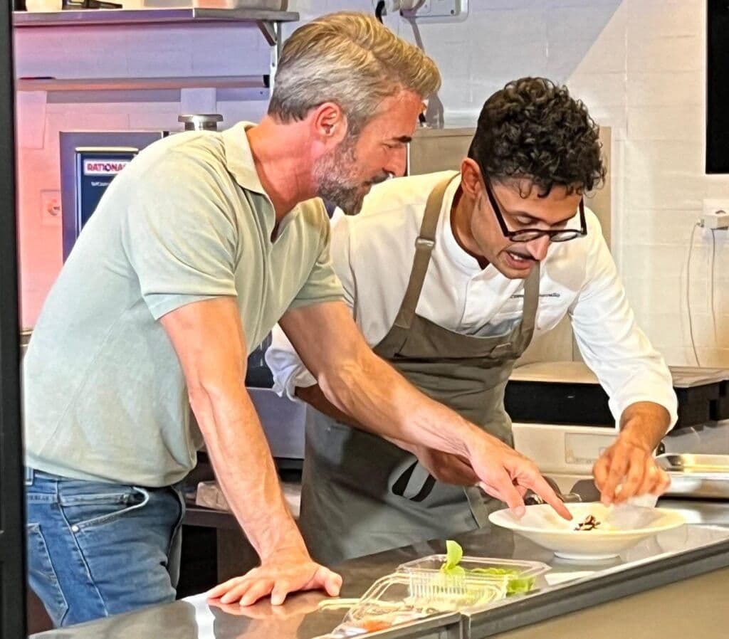 Flavio Montrucchio et un chef cuisinier préparant un plat, illustrant l'innovation culinaire et la symphonie de saveurs de Milazzo.