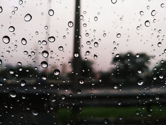 Gotas de chuva em uma janela, com o fundo borrado de uma rua, representando o impacto da chuva no dia a dia.