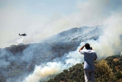Hélicoptère bombardier d'eau luttant contre un incendie de forêt en Turquie, avec des collines fumantes en arrière-plan.