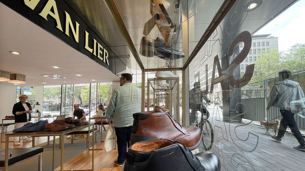 Interior view of a Van Lier shoe store with shoes on display and a 'SALE' sign reflected in the window, hinting at the brand's financial struggles.