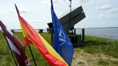 Latvian, Spanish, and NATO flags fluttering in front of an air defense missile system on a field, representing allied unity and air defense capabilities.