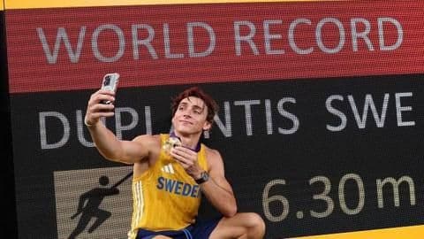 Le perchiste Armand Duplantis prenant un selfie devant le tableau des records du monde, symbolisant l'attention médiatique.