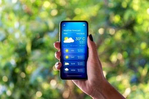 Main tenant un smartphone affichant une application de prévisions météorologiques avec un ciel ensoleillé et des températures.